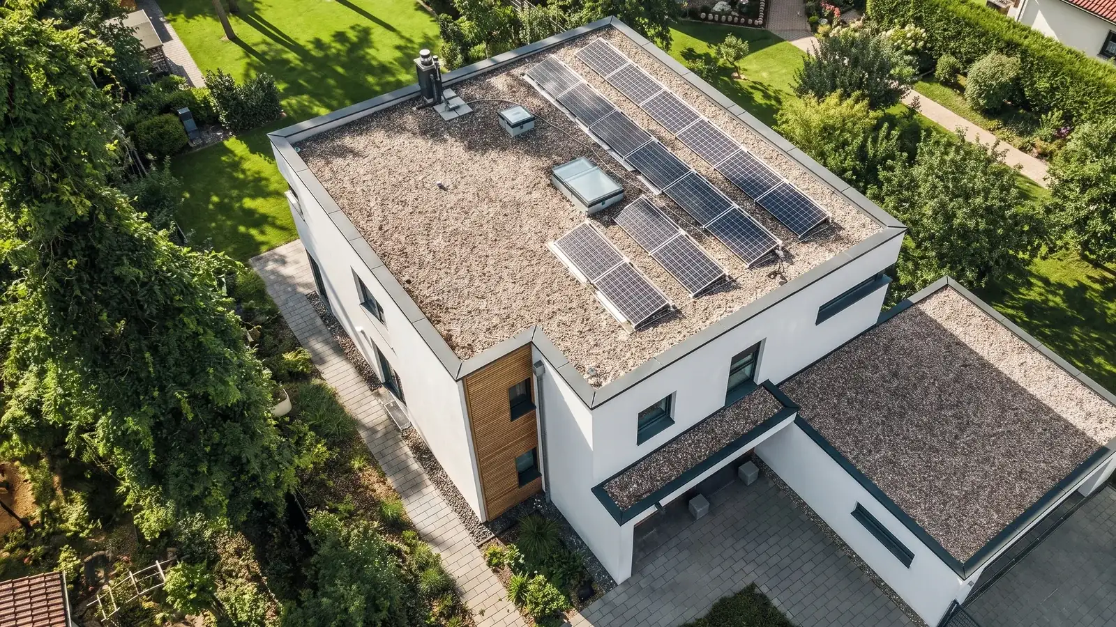 Luftbild eines modernen Wohnhauses mit Flachdach: Kiesauflast auf der Abdichtung, aufgeständerte Solarmodule, ein flaches Dachfenster und eine umlaufende Attika mit dunkler Blechkante, daneben das Flachdach der Garage